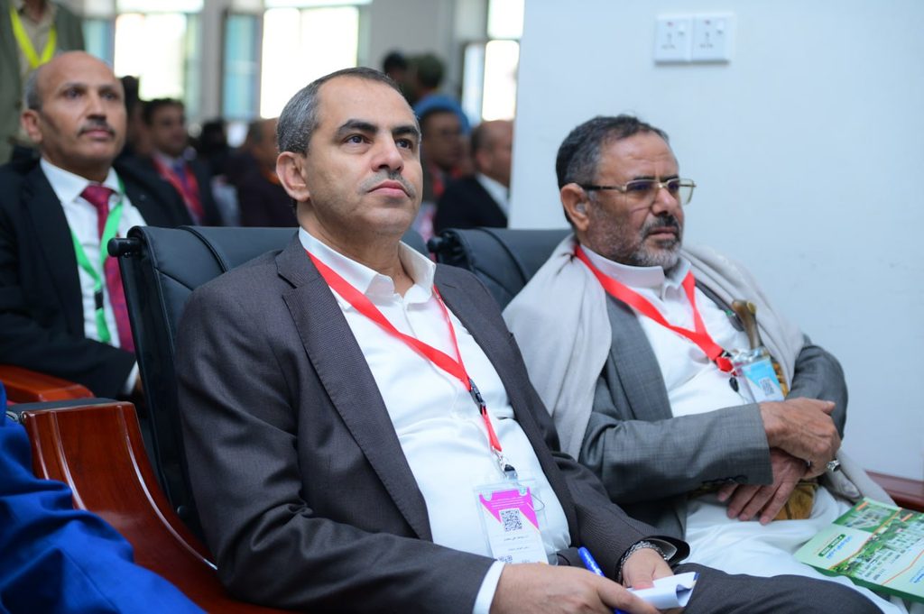 IMG_20260208_072709_620 - جامعة 21 سبتمبر Two men in suits seated in a conference room wearing red lanyards and badges in front rows of an event.