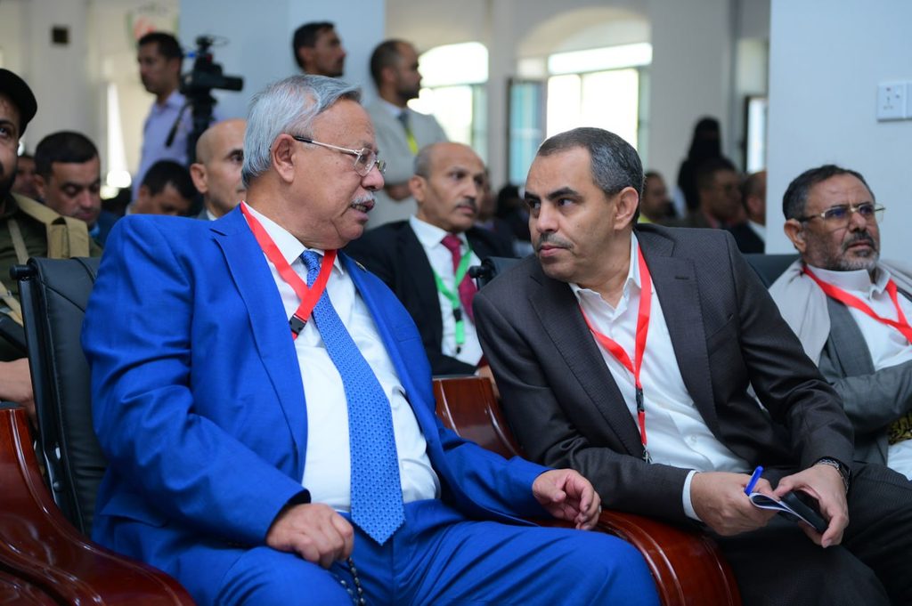 IMG_20260208_072709_651 - جامعة 21 سبتمبر Two men in suits wearing red lanyards sit side by side in a conference hall, engaged in conversation while others listen in the background.