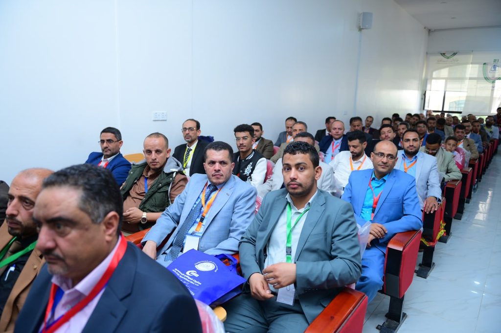 IMG_20260208_072720_568 - جامعة 21 سبتمبر Group of men in business attire seated in a conference room, wearing name badges and lanyards.