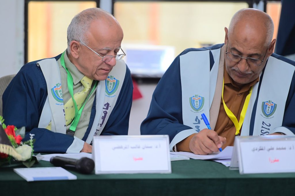 IMG_20260208_082702_076 - جامعة 21 سبتمبر Two older men in ceremonial robes sit at a table signing documents during a formal event, with nameplates and badges on their vests visible on the table edge.