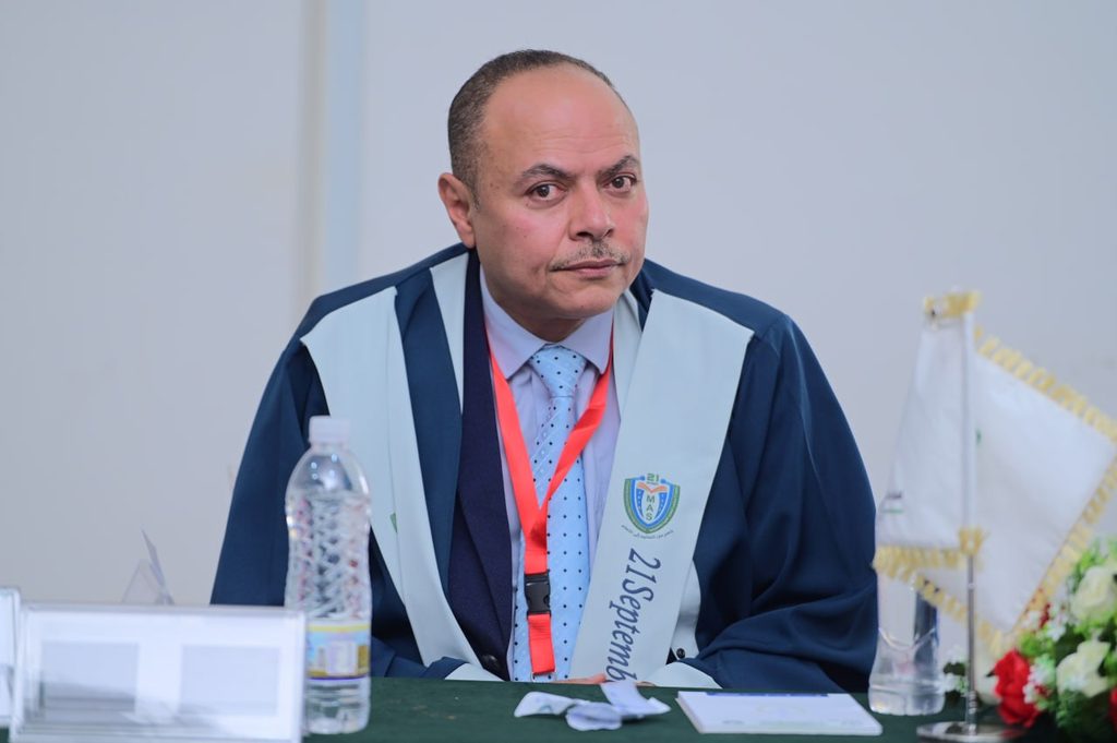 IMG_20260208_082702_645 - جامعة 21 سبتمبر Middle-aged man in academic gown and light-blue sash seated at a table during a ceremony, with a water bottle nearby.