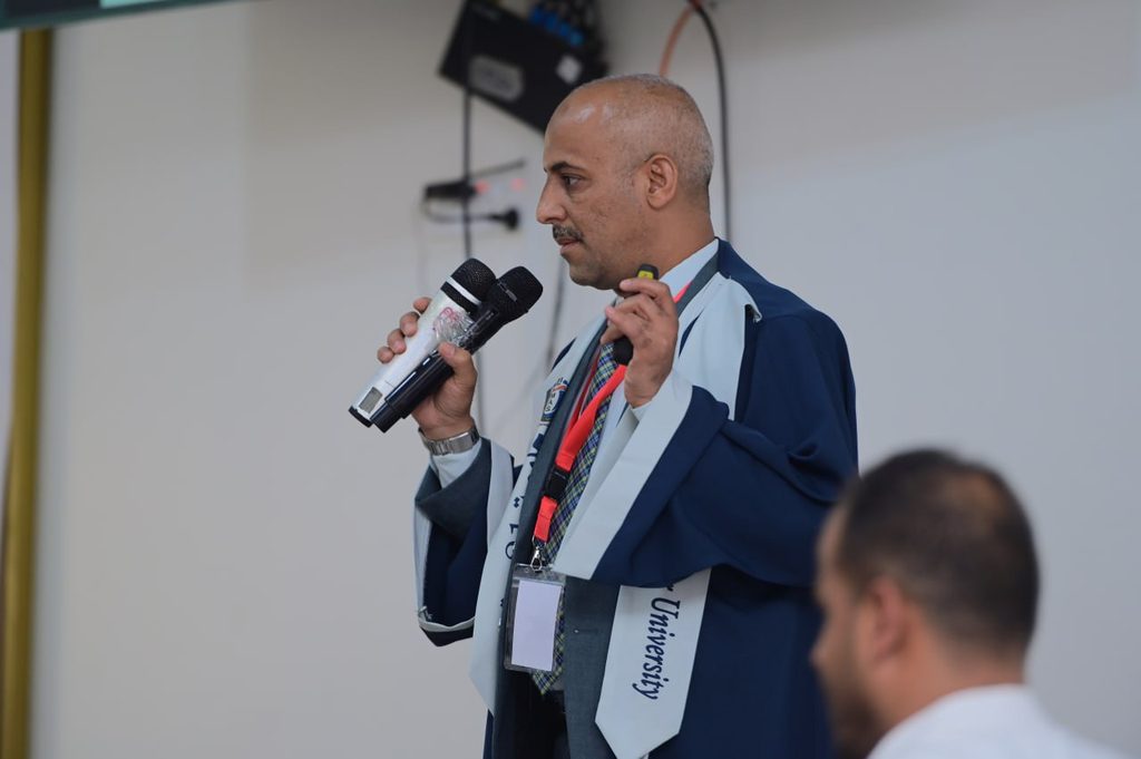 IMG_20260208_082702_697 - جامعة 21 سبتمبر Man in academic gown speaking into two wireless microphones on stage, wearing lanyard and badge on a conference backdrop.