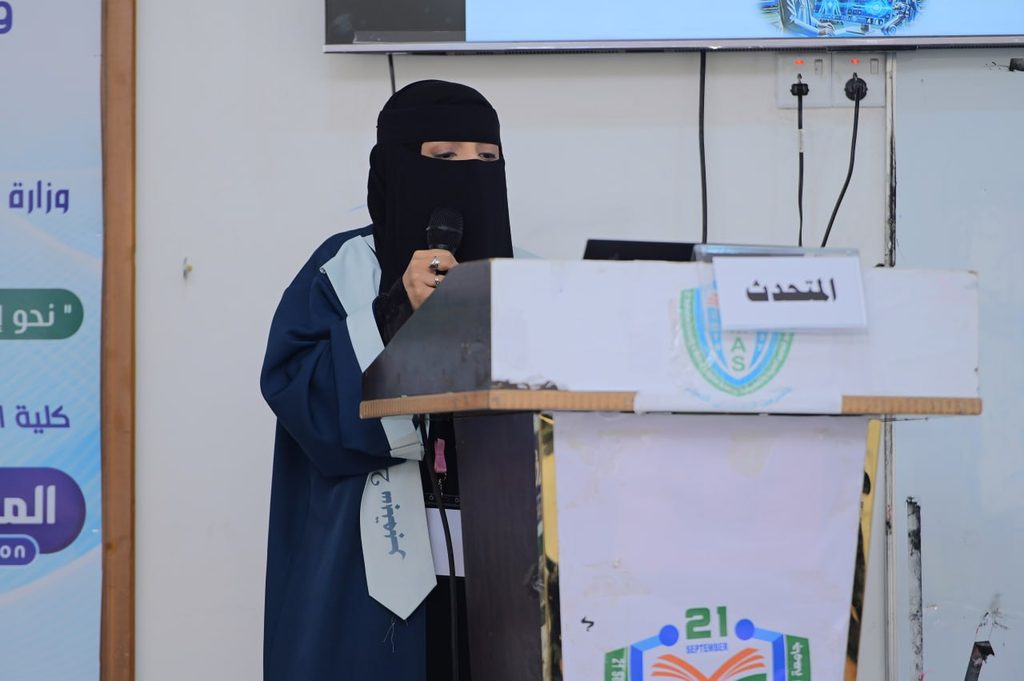 IMG_20260208_082710_032 - جامعة 21 سبتمبر Woman wearing a black niqab and blue gown speaks into a handheld microphone at a podium.