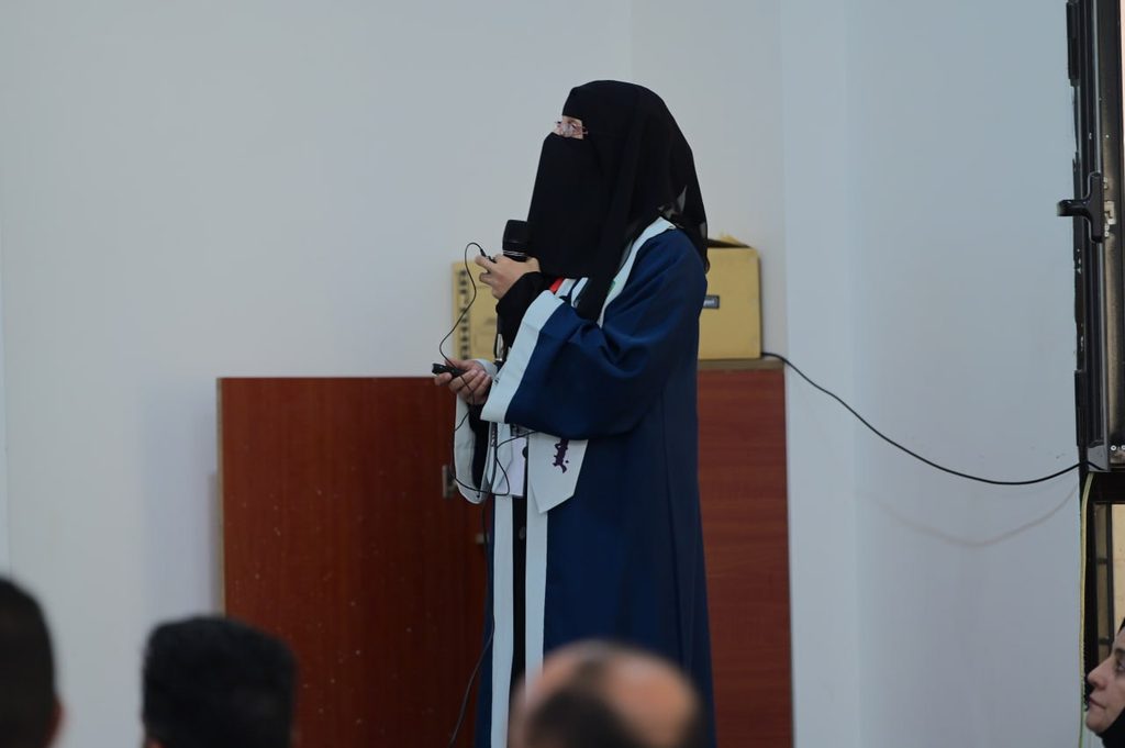 IMG_20260208_082727_008 - جامعة 21 سبتمبر Woman in a blue graduation gown and black hijab speaks into a handheld microphone at an indoor event, addressing an audience.