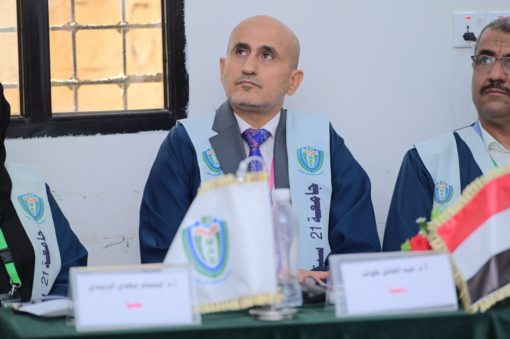 IMG_20260208_082727_101 - جامعة 21 سبتمبر Bald man in a blue graduation gown and light blue stole sits at a table during a formal ceremony, looking upward.
