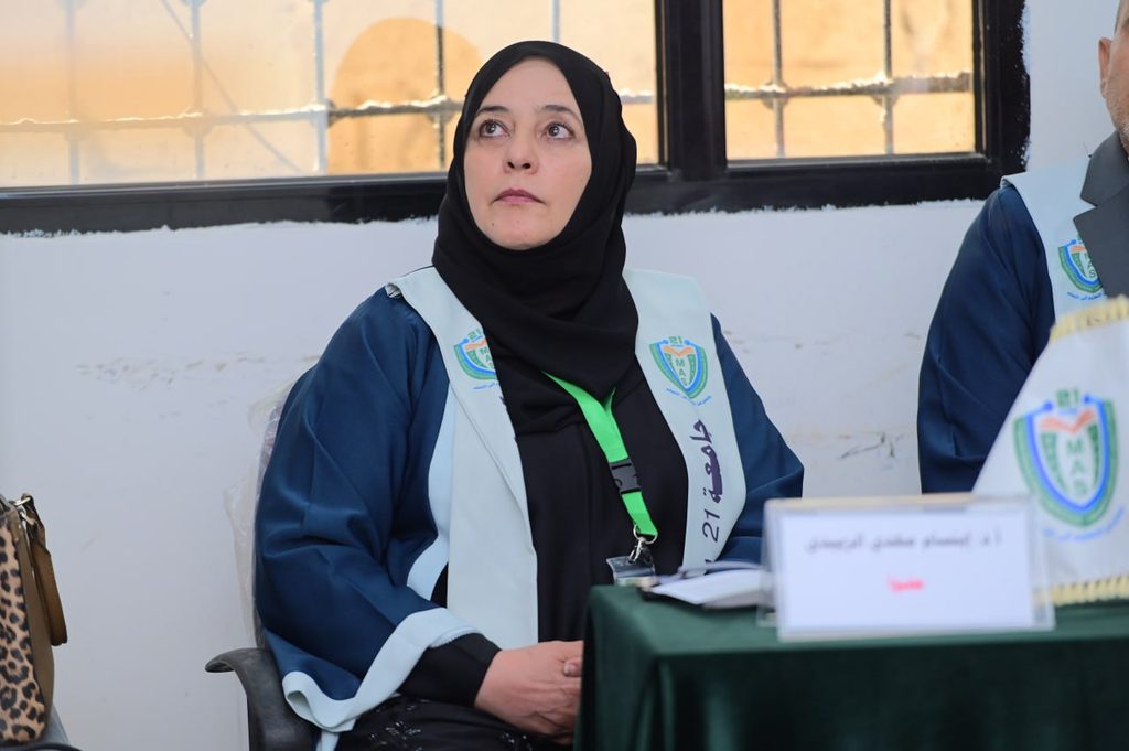 IMG_20260208_082727_132 - جامعة 21 سبتمبر Panelist wearing a black hijab and blue robe sits at a table with a nameplate during a formal meeting.
