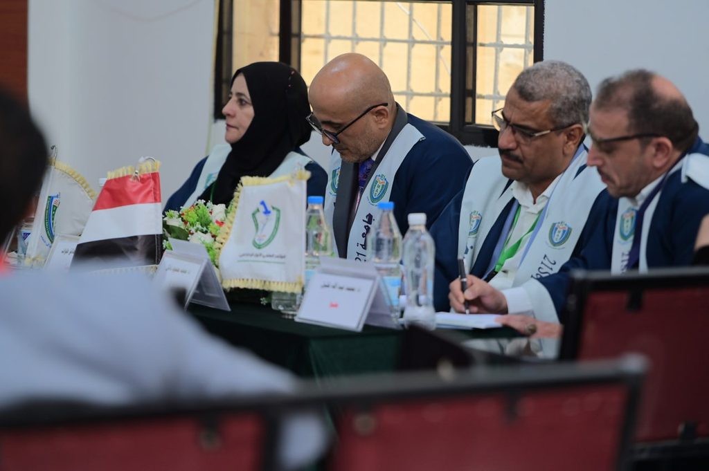 IMG_20260208_082727_302 - جامعة 21 سبتمبر Group of officials in blue attire with light blue sashes seated at a conference table, flag and flowers in front.
