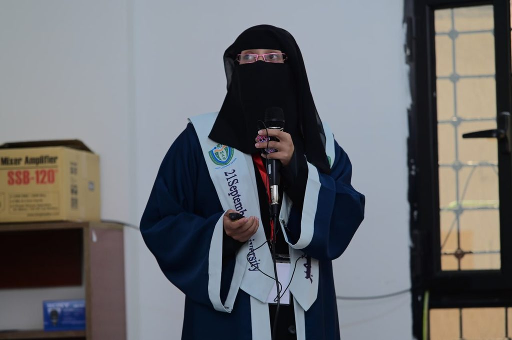 IMG_20260208_082727_720 - جامعة 21 سبتمبر Woman in a blue graduation gown and black niqab speaks into a microphone at an indoor ceremony, sash across chest