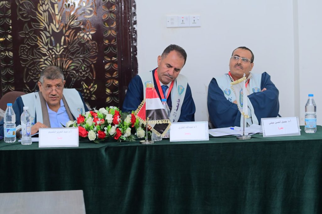 IMG_20260208_082735_088 - جامعة 21 سبتمبر Three men in blue robes sit behind a green-draped table with nameplates, flowers, and small flags at a formal panel event.