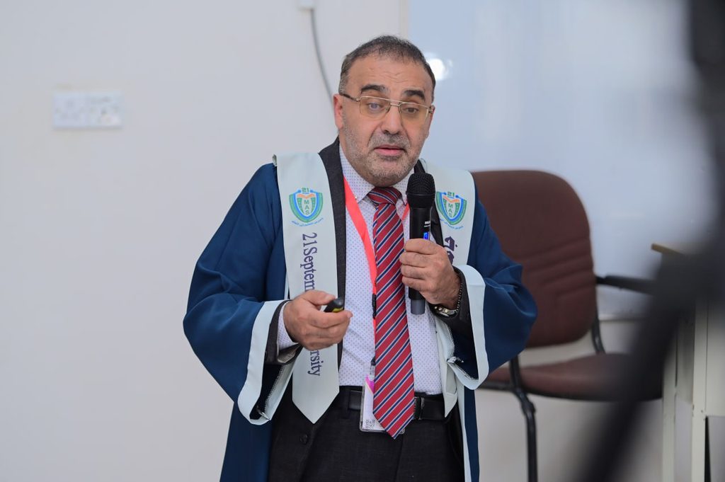 IMG_20260208_082917_800 - جامعة 21 سبتمبر Middle-aged man in blue academic robes speaks into a handheld microphone during a presentation, holding a remote.