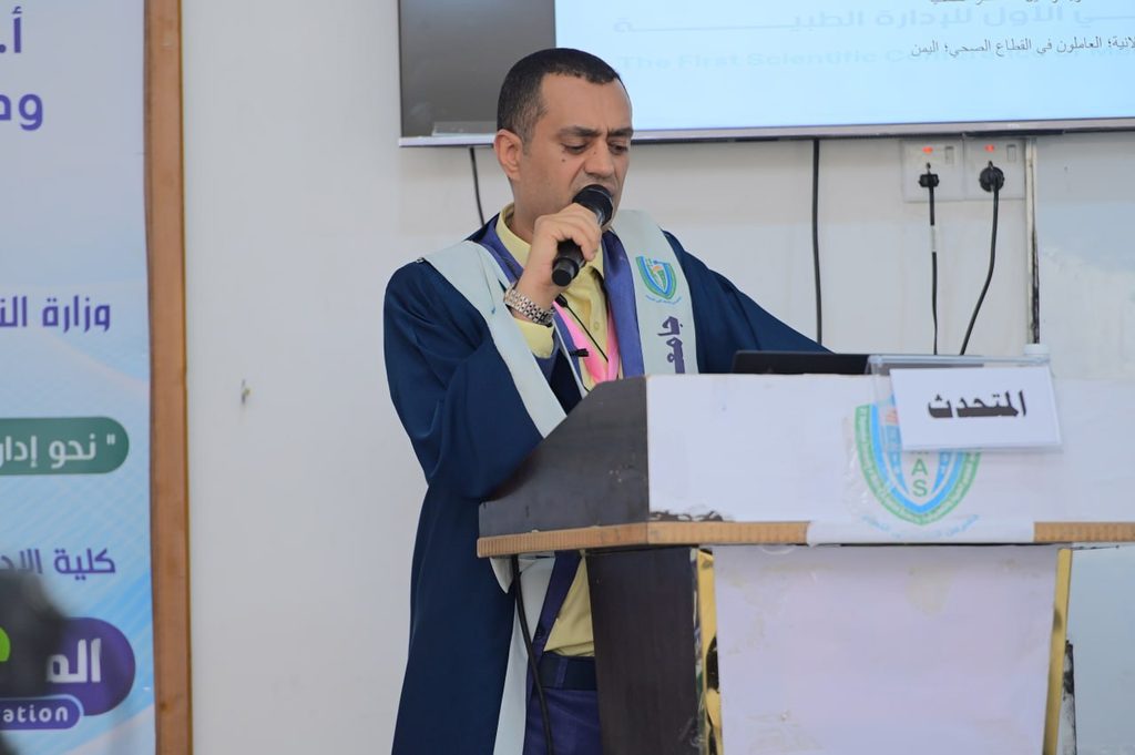IMG_20260208_082917_997 - جامعة 21 سبتمبر Man in a blue graduation gown speaks into a handheld microphone at a podium during a formal event, with banners in the background.