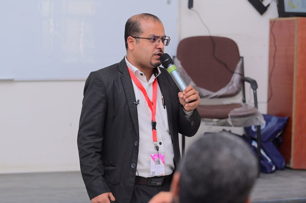 IMG_20260208_084313_459 - جامعة 21 سبتمبر Male speaker in a dark suit and white shirt, wearing a red lanyard, speaks into a handheld microphone at a conference.