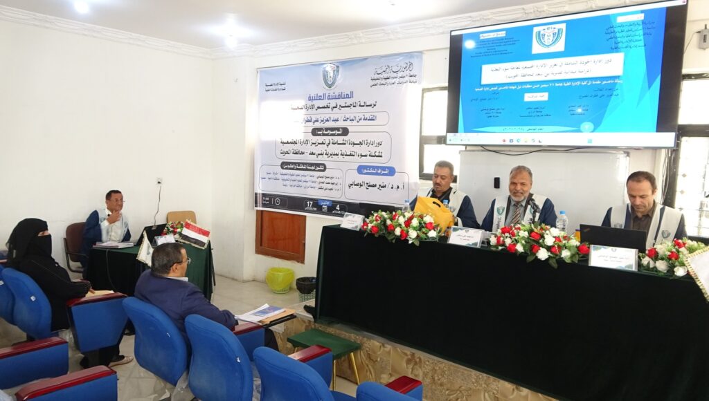 Panel discussion at a conference: four men sit behind a long table with flowers; banner and large screen with Arabic text behind them; audience seated in blue chairs