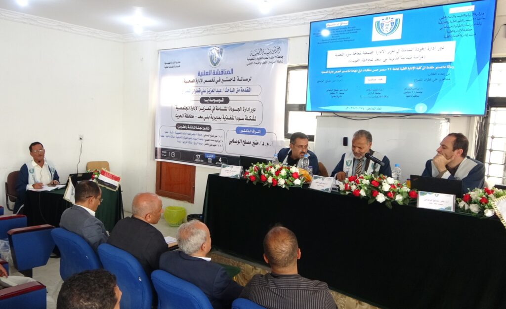 Panel of men seated at a long black-draped table with flower garlands, microphones, and a large blue banner behind them (Arabic text).