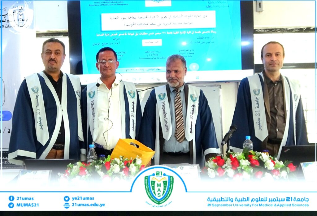 Four men in blue-and-white academic gowns stand behind a conference table at a university ceremony, with a blue presentation screen and floral arrangements in front.