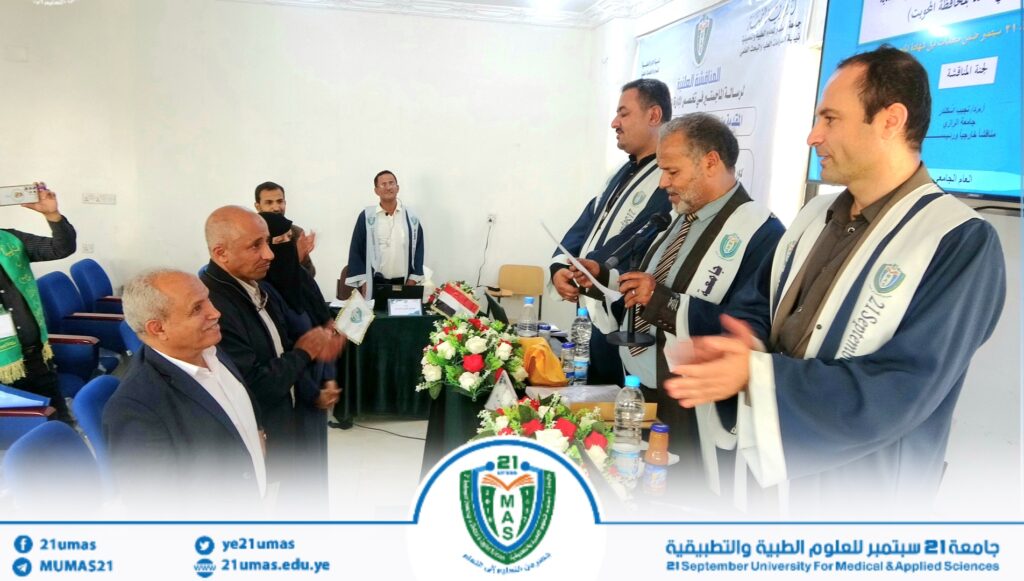 Panel of men in ceremonial robes speak at a formal event, with a flower‑decorated table and a university crest in the foreground and a blue banner in Arabic in the background.