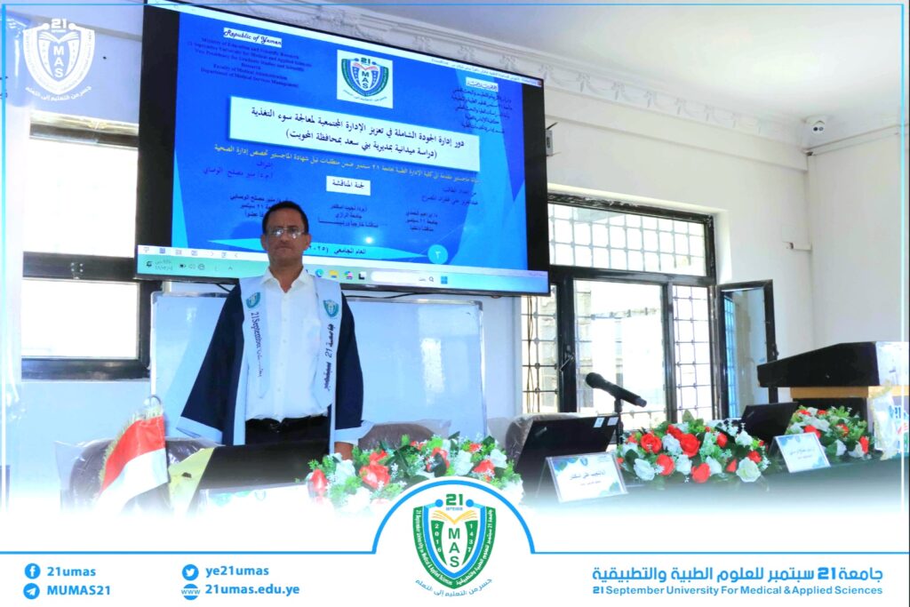 Speaker in ceremonial robes stands at a podium during a university presentation, with a large blue slide and MAS emblem on screen behind him and floral decorations in front.
