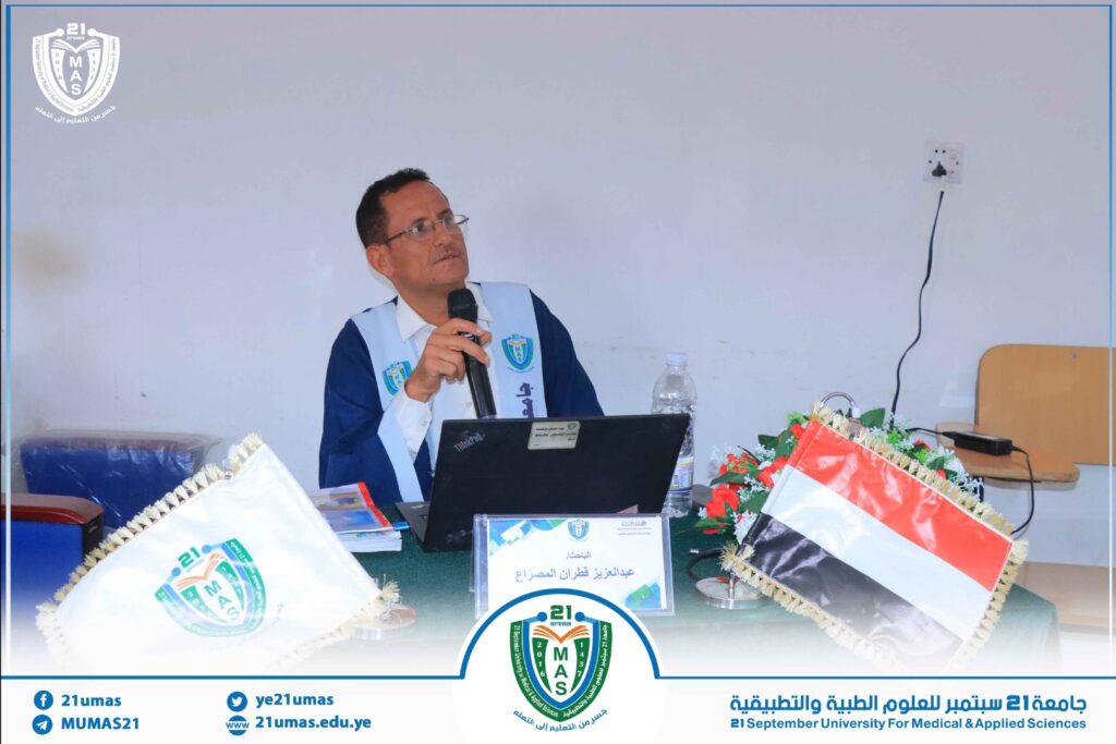 Man in a blue-and-white vest speaks into a microphone at a desk with a laptop, flags, and a university emblem in the background.