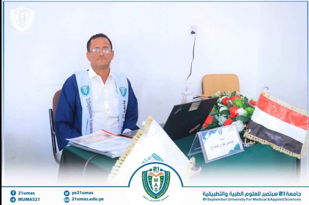Man in a blue academic gown seated at a desk with a laptop, MAS logo banner, and a flag, at a university event.