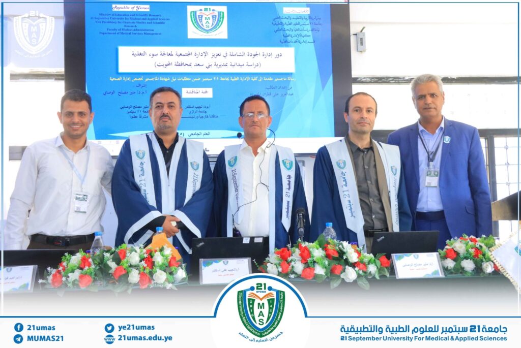 Five men pose at a university ceremony; three wear blue robes with white sashes, backdrop displays Arabic text and university logos.