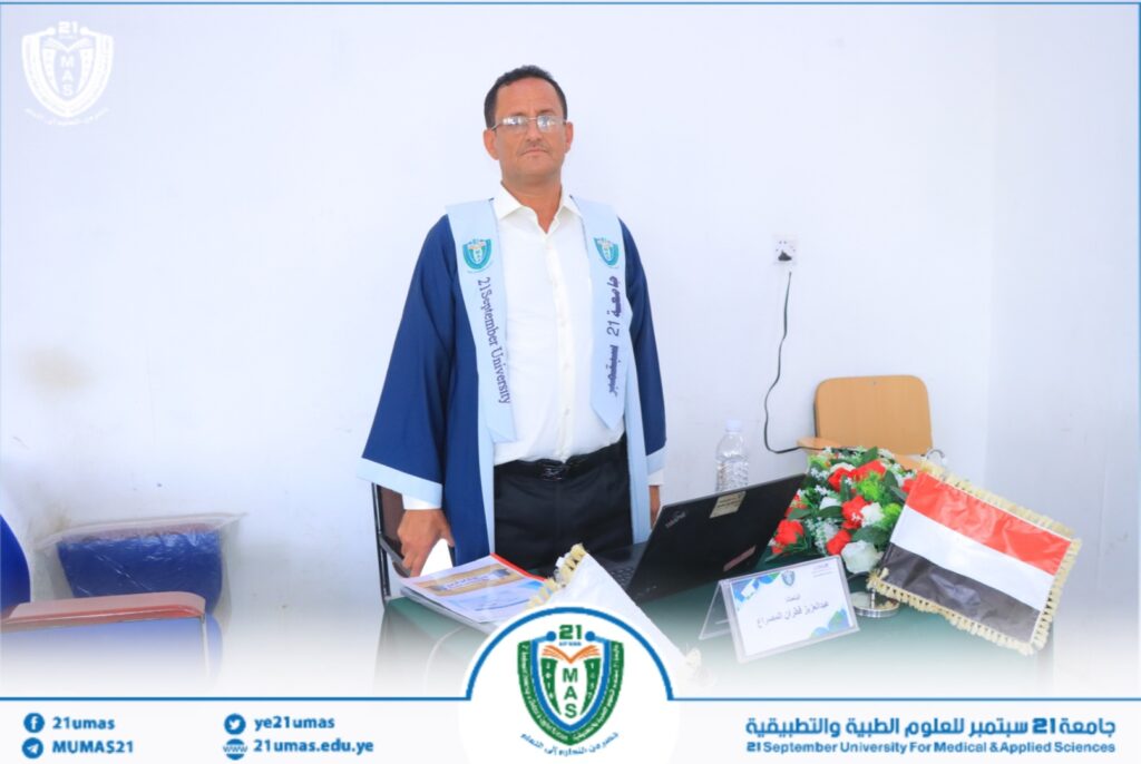 Man in a blue graduation gown and white shirt stands behind a desk with a laptop, floral arrangement, and a small Yemeni flag on the table, in a formal university setting.