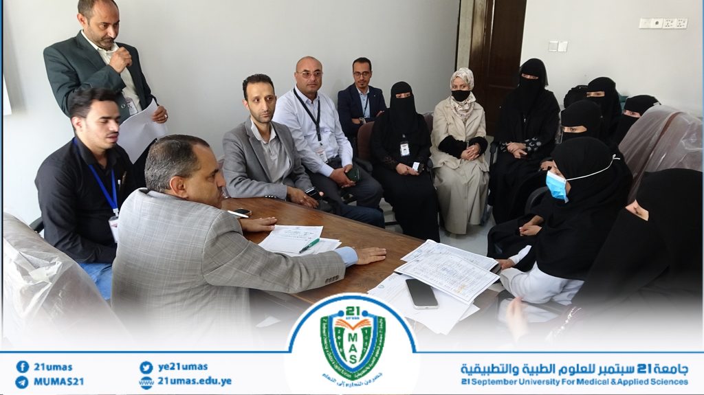 Group of professionals seated around a table in a meeting, a man stands with papers while others listen, some women in hijabs and masks present.