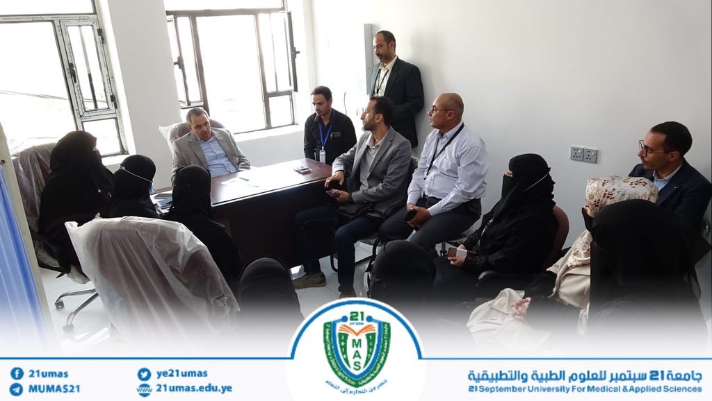 A group of professionals, including women in black abayas and men in suits, sit around a conference table in a bright office; a man stands by the wall.
