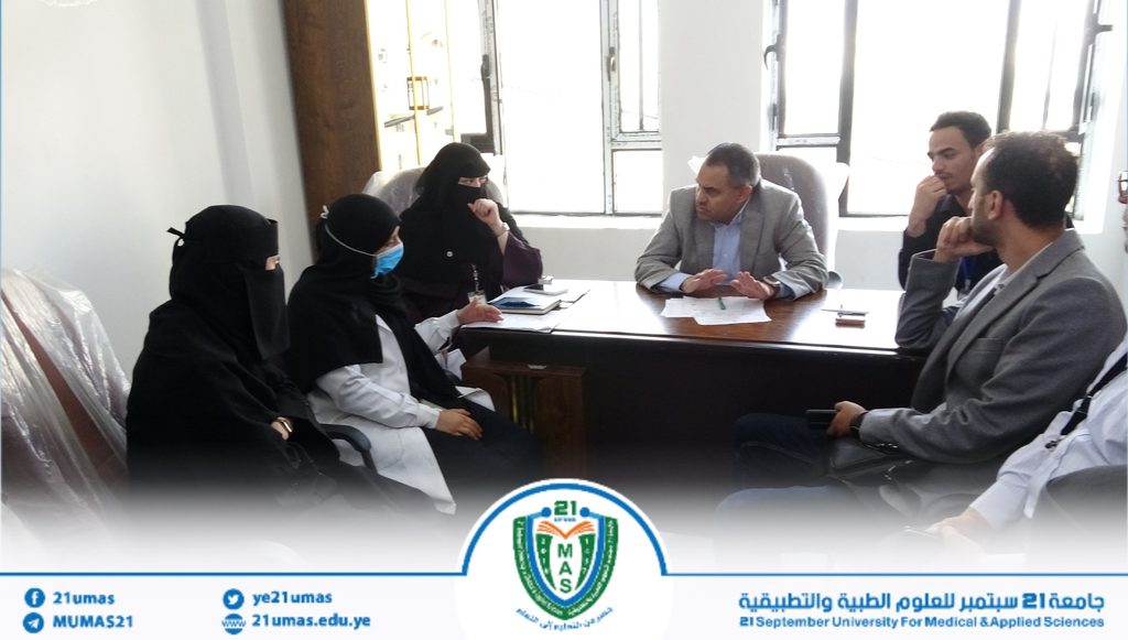 Group of people in a formal meeting around a desk in a bright office; a university banner with a logo is visible at the bottom center.