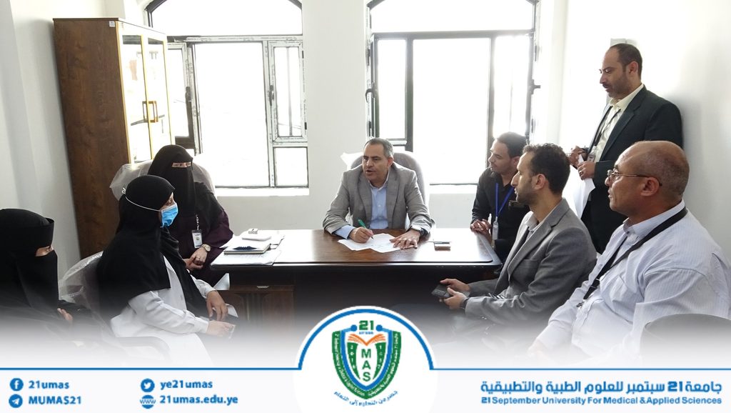 Group of professionals in a meeting around a wooden desk; man in a gray blazer speaks while others listen, including women in hijabs, in a bright room with a university banner.
