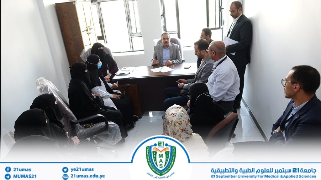 Group of professionals in a small office meeting around a desk; a man in a gray blazer speaks as others listen, including several women wearing hijabs.