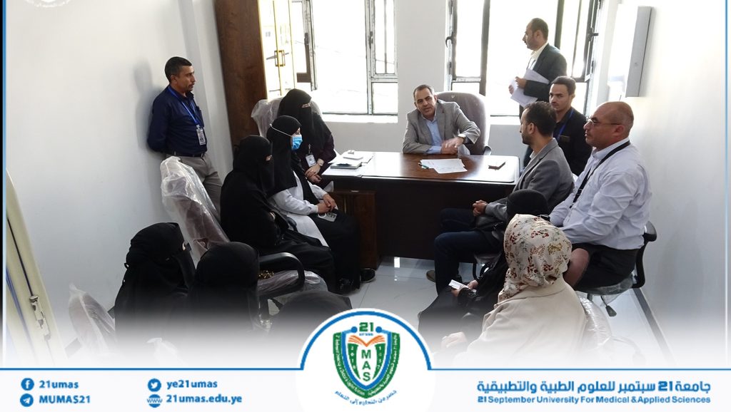 Group of professionals in a meeting around a desk in a bright office, some women wearing hijabs, one man in a suit extending a handshake across the table.