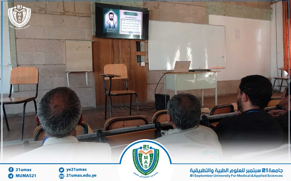 Lecture in progress at 21 September University: a TV screen displays Arabic text, with a whiteboard, laptop on a desk, and students facing the front.