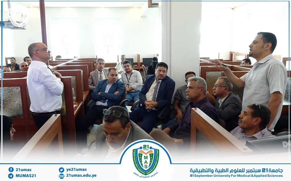 Lecture hall scene: a presenter on the right speaks to a group of men in suits seated and listening attentively.