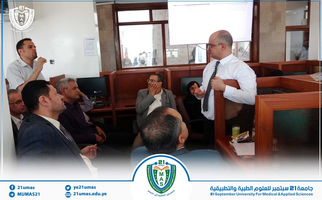 Man in a white shirt and tie speaking to seated colleagues in a university meeting room, MAS logo visible on a banner at the front.