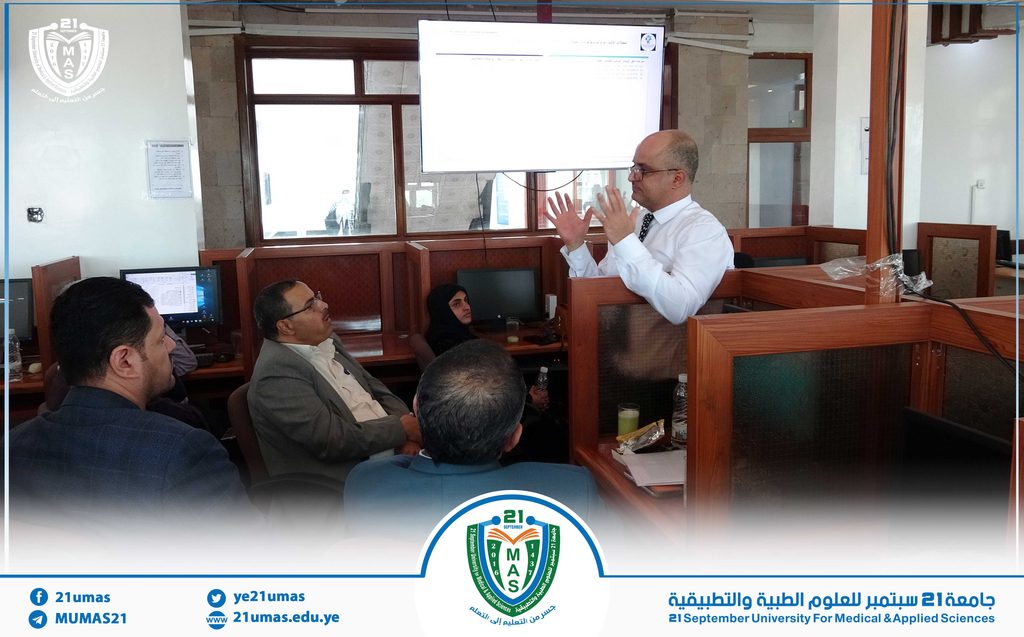 Instructor presenting at a university lecture, gesturing with hands while seated attendees listen nearby in a wood-paneled, computer-filled room.