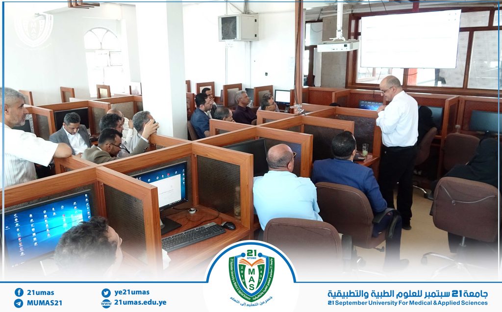 Lecture in a university IT lab: a man in a white shirt presents to seated professionals at computer cubicles with a projector screen in the background.