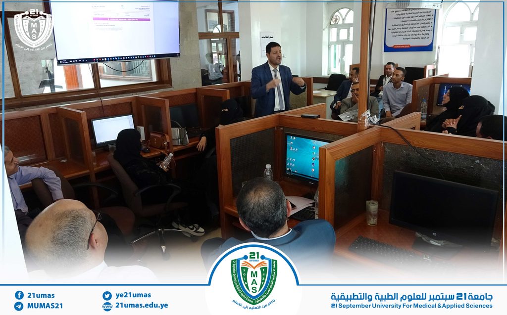 Male lecturer in a blue suit presenting to a seminar audience in a university computer lab with cubicles and monitors visible