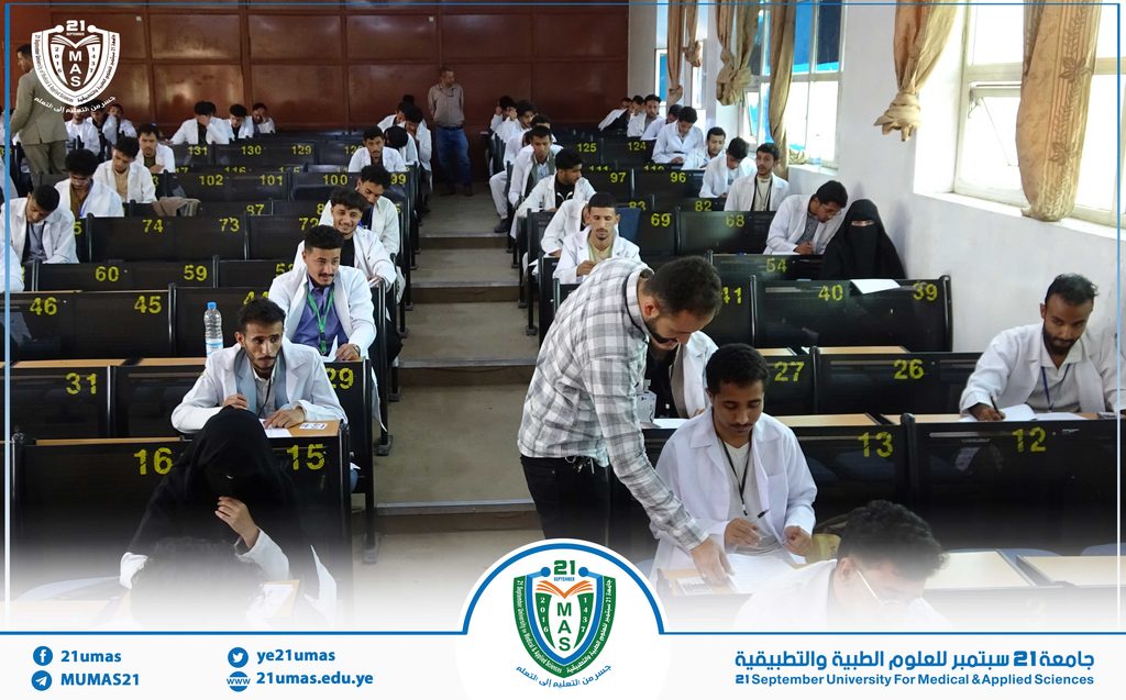 Students in white coats taking an exam in a university lecture hall; desks are numbered and a staff member assists at the front.