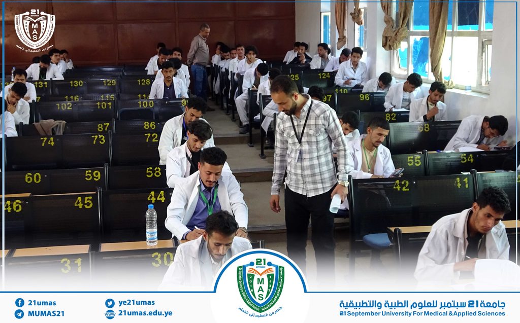 Group of students in white lab coats seated in a lecture hall with numbered seats, university logo watermark on the left and event banner at the bottom.