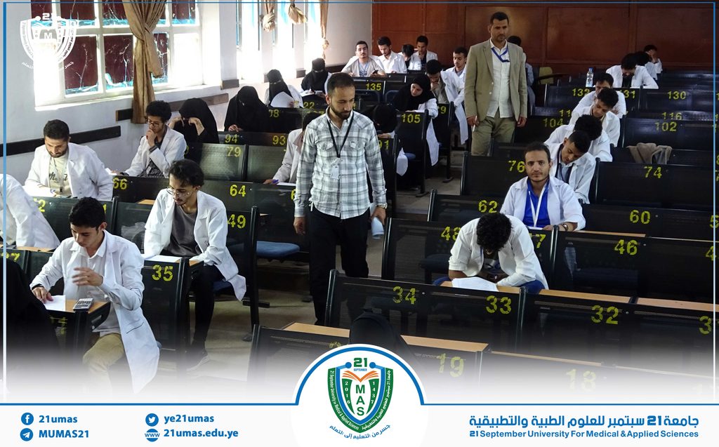 Students in white coats sit in a lecture hall, taking notes while a man in a plaid shirt stands at center; yellow seat numbers visible on chairs and a university logo appears at bottom.