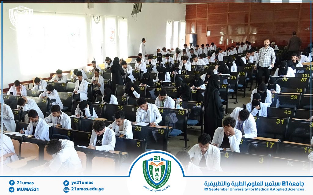 Lecture hall full of students in white coats taking notes while instructors walk between desks in a university setting.