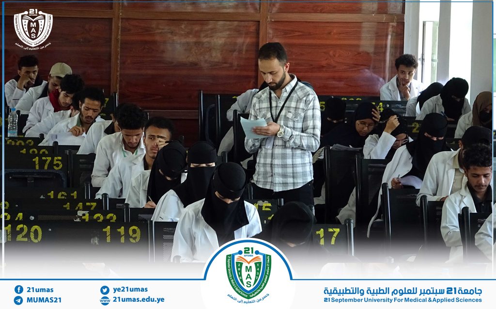 Students in white lab coats take notes in a lecture hall while a man stands reading from papers; university event logo visible in foreground.