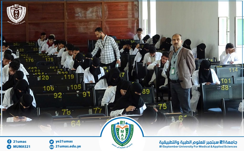 Exam hall at 21 September University; students in white coats (many women in hijabs) seated at numbered desks while a man in a plaid shirt walks through.