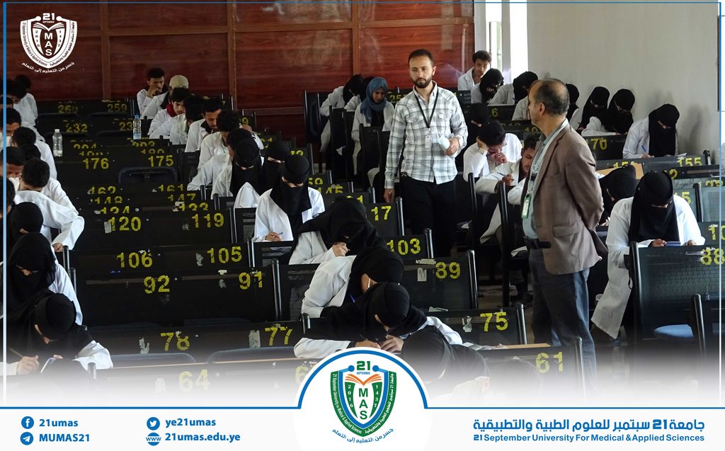 Exam hall with students in white coats taking notes as a man in a checkered shirt walks among desks; university emblem visible on the banner at the front.