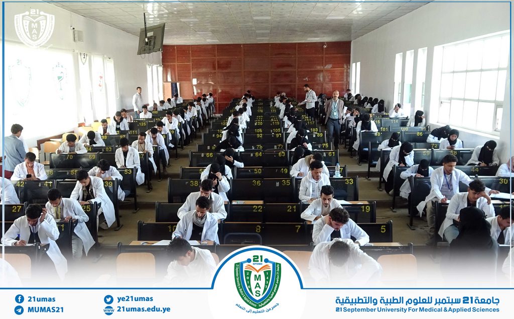 Students in white lab coats sit at numbered desks taking exams in a large hall; classroom full of test takers with a university logo visible at the bottom.