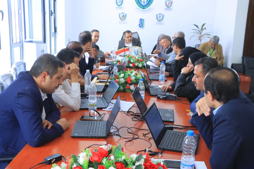 Group of professionals around a long conference table with laptops, bottled water, and a floral centerpiece during a meeting.