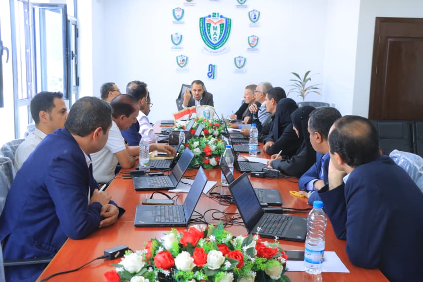 Group of professionals around a long conference table with laptops, bottled water, and a floral centerpiece, in a room with a shield logo on the wall behind them.