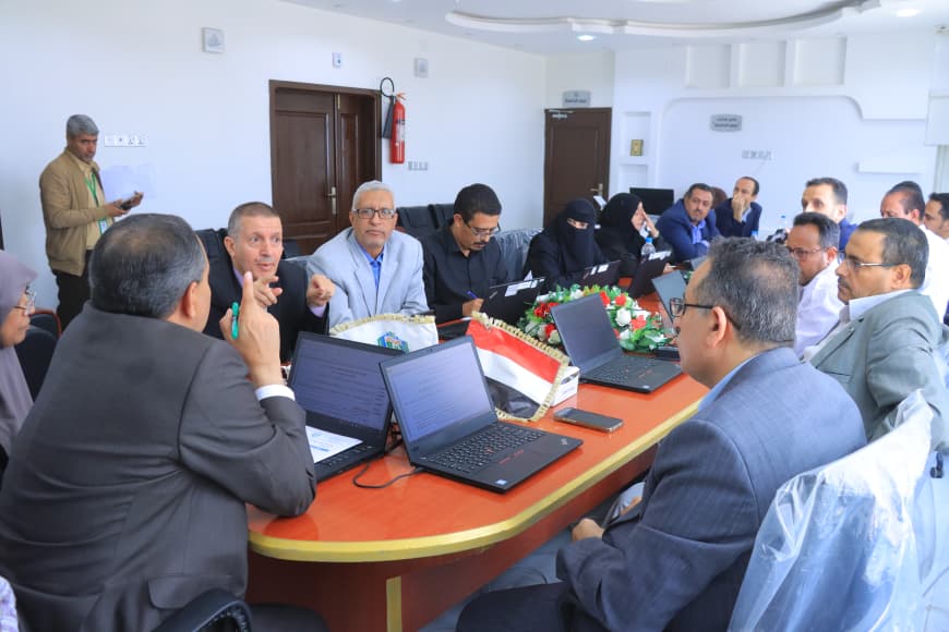 A diverse group of professionals seated around a long conference table with laptops, discussing ideas; a small national flag centerpiece sits in front of the group.