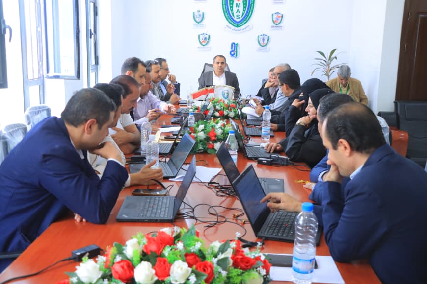 Group of professionals in a conference room seated around a long orange table with open laptops and bottled water close to a floral centerpiece on display behind them.