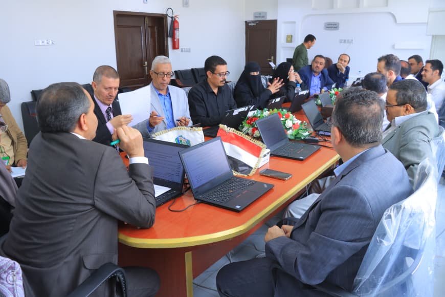 Business meeting with numerous professionals around a long table, laptops open and flags in the center.
